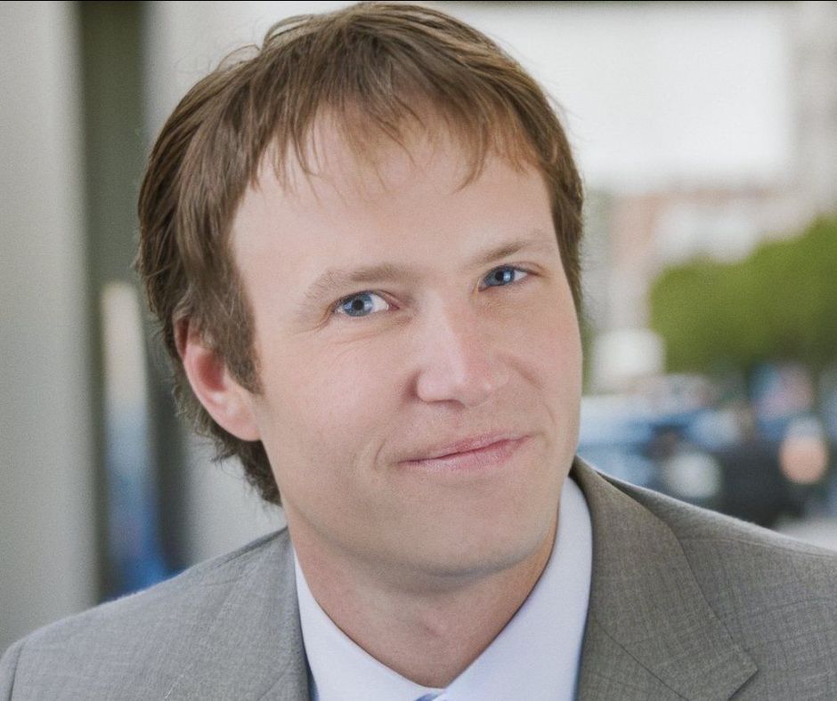 Man in gray suit smiles, light brown hair, blue eyes, blurred city background.
