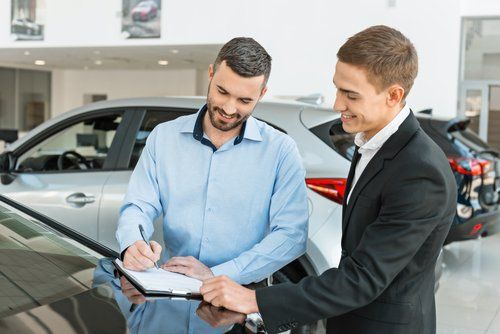 A man is signing a document in front of a car while another man looks on.