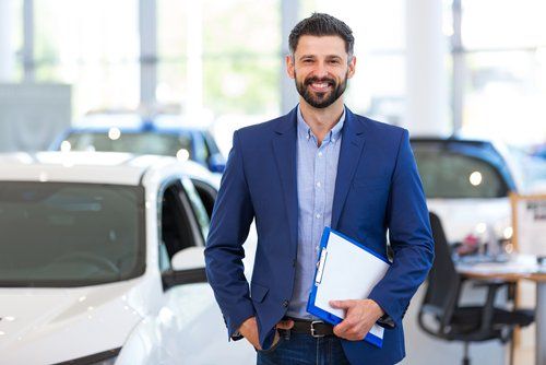 A man in a suit is holding a clipboard in front of a car in a showroom.