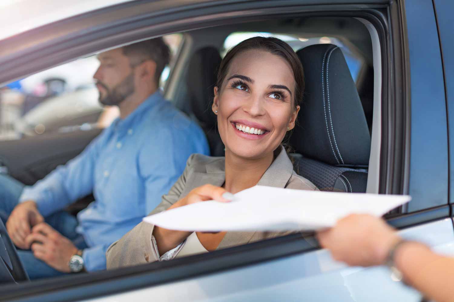 Woman in car smiles, receiving document from person outside. Man in background. Sunny daytime.