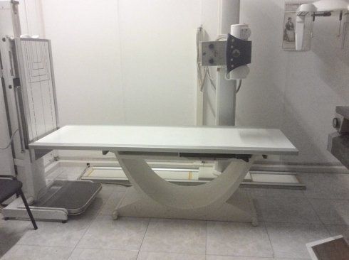 Tavolo radiografico ospedaliero in una sala clinica, di colore bianco con base di supporto curva e apparecchiature circostanti.