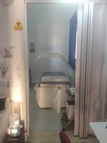 Piccola stanza clinica con lettino per visite, lavandino, tenda per la privacy e cartello di avvertimento sulle radiazioni.