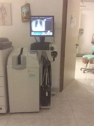 Sala per visite mediche con un'unità radiografica portatile e un monitor accanto a un dispositivo bianco sul pavimento.