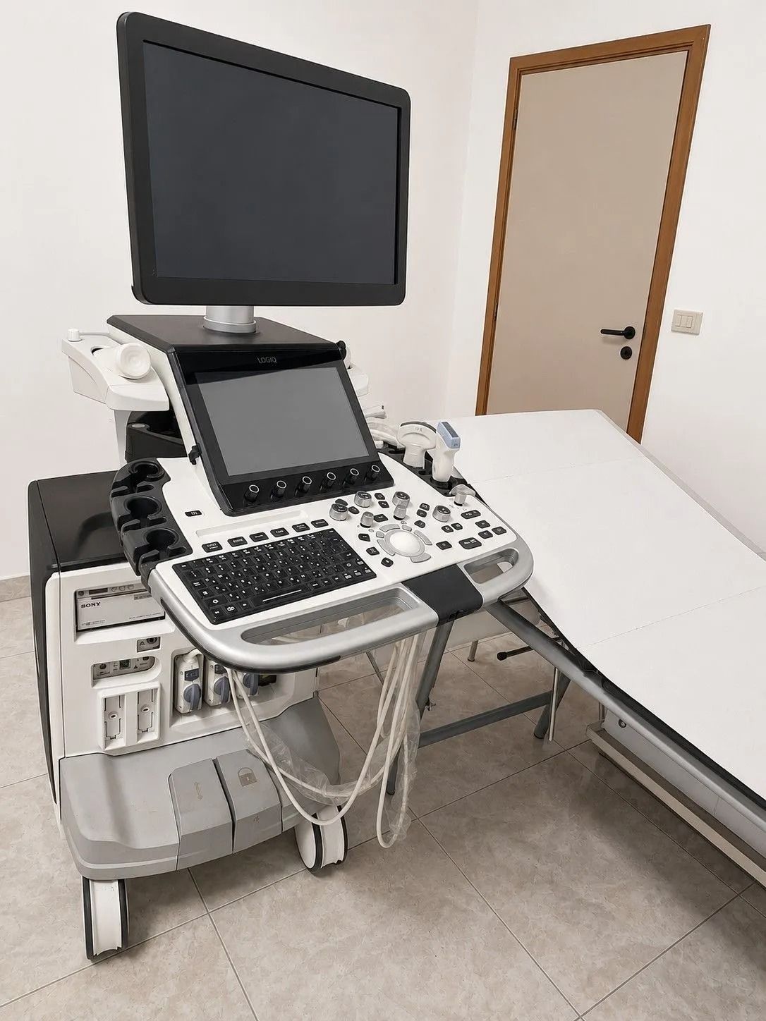 Apparecchio ecografico medicale accanto a un lettino da visita in una stanza clinica con porta e monitor a parete.