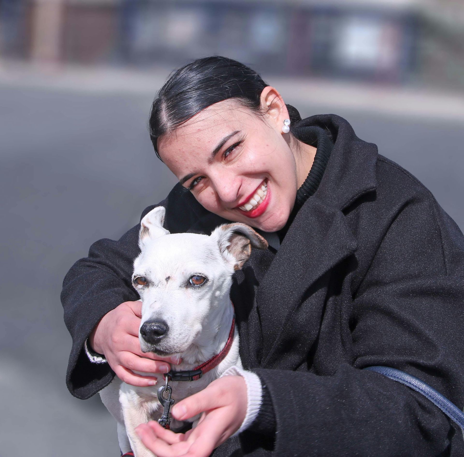 Person in a black coat smiling and hugging a white dog outdoors