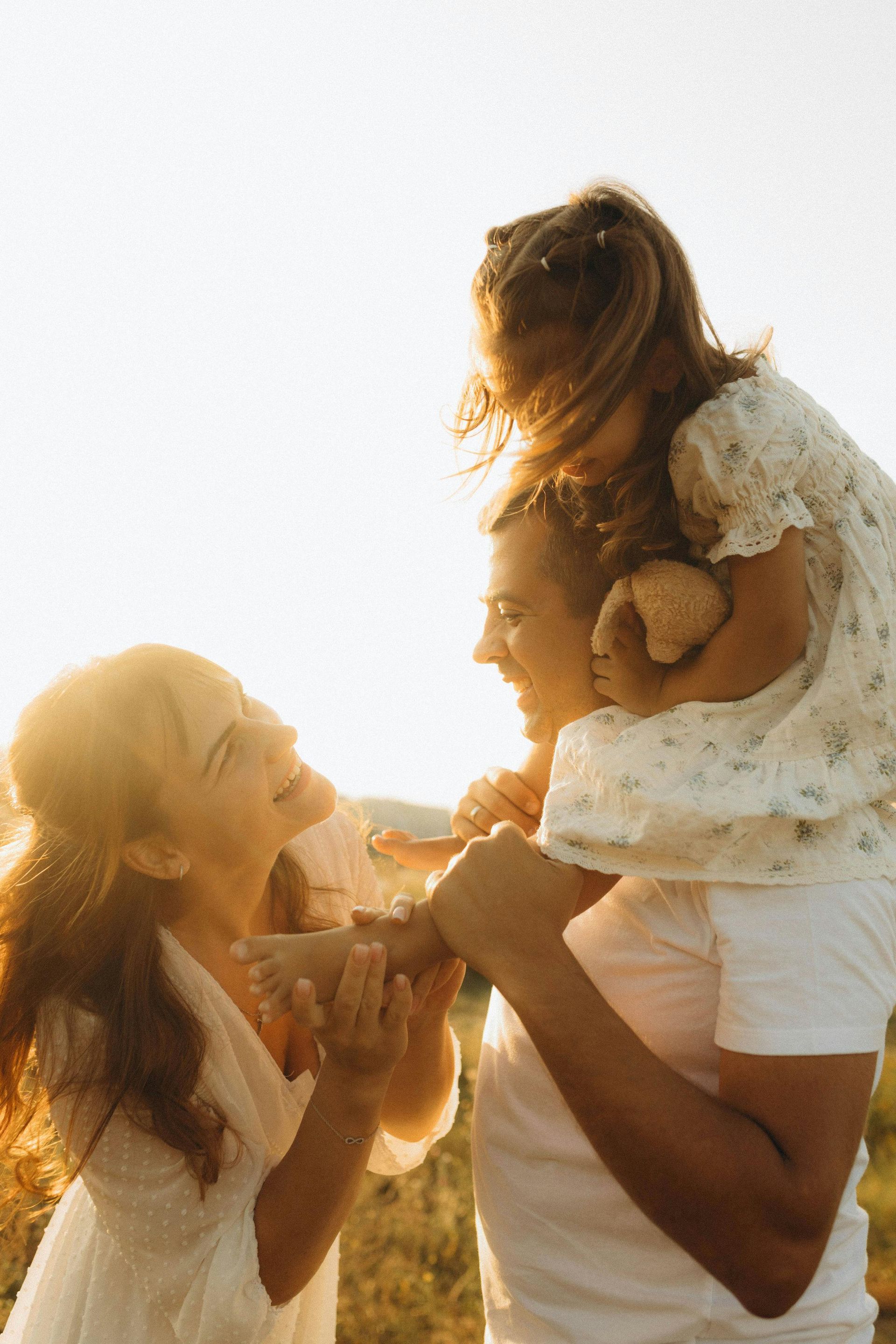 Two adults lifting a child outdoors in warm sunlight, smiling and facing each other.