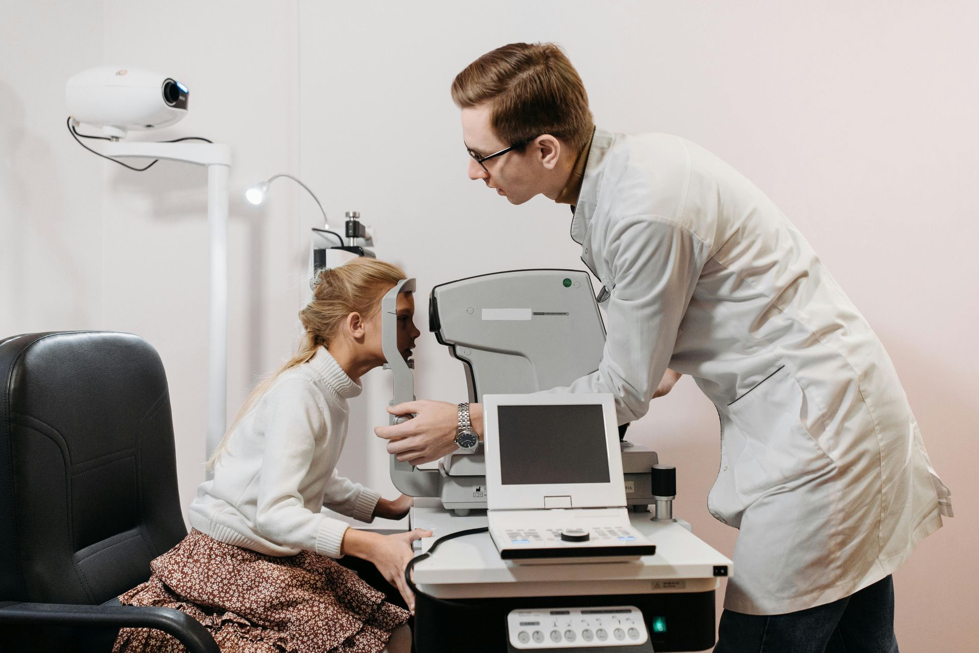 Eye doctor examining a child in an exam room with diagnostic equipment on a desk