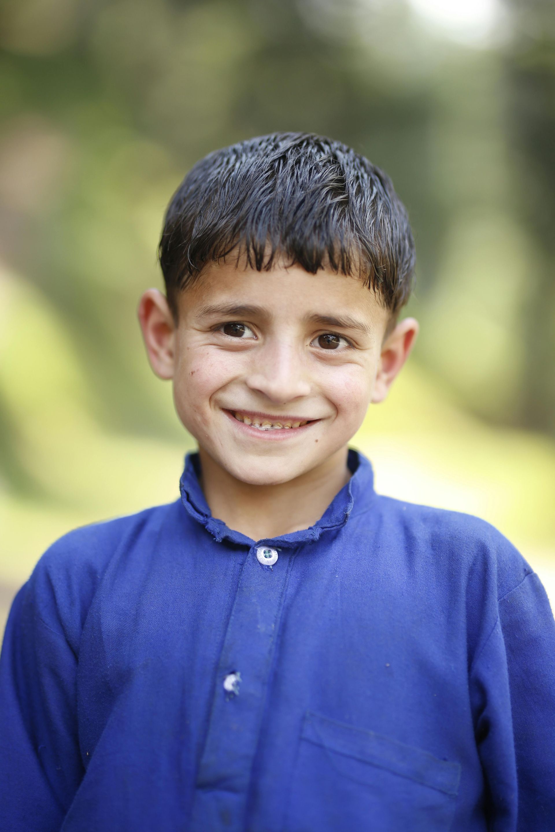 Smiling child in a blue shirt standing outdoors with a blurred green background