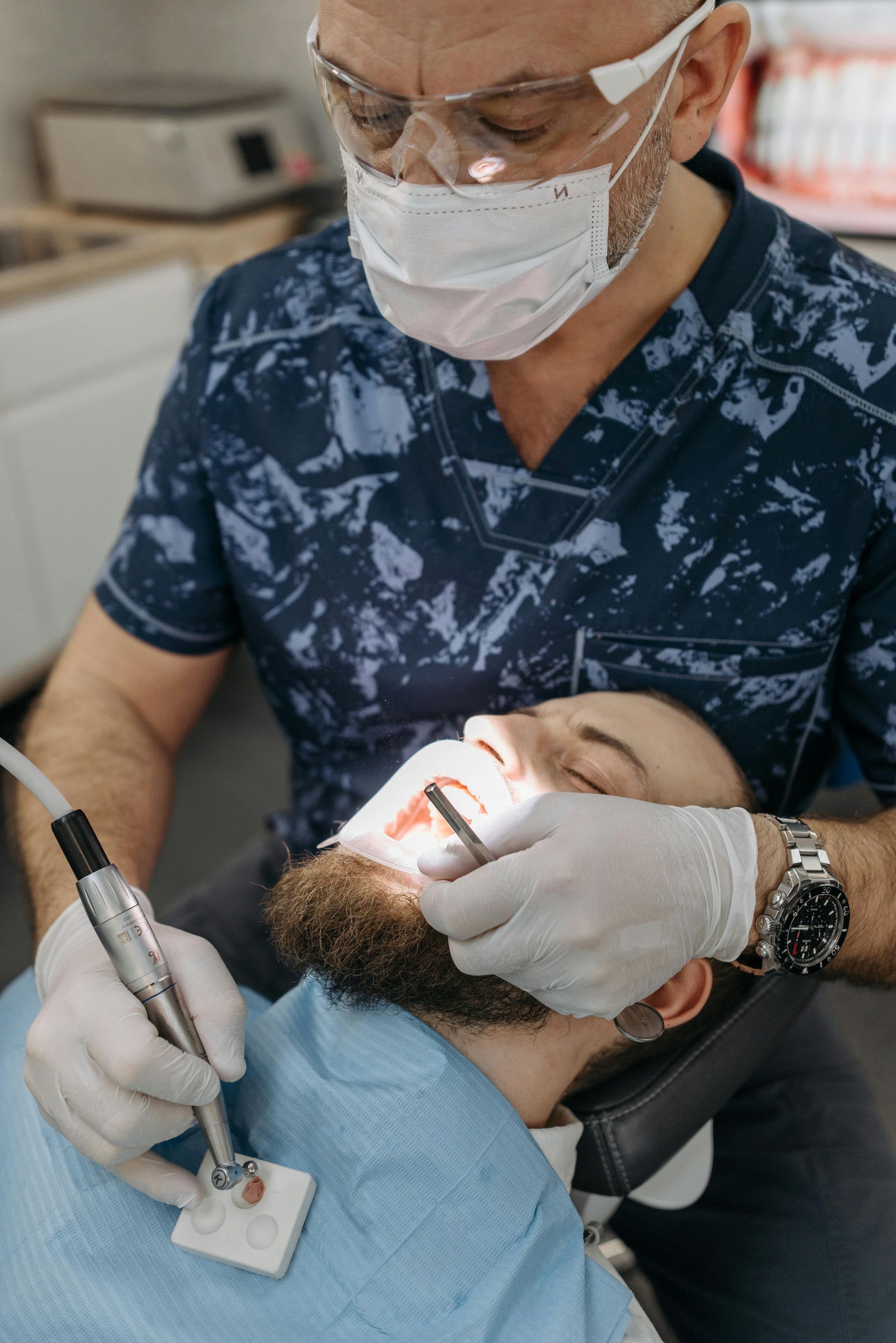 Dentist wearing gloves and mask examines a patient in a dental chair with a drill and mouth light.