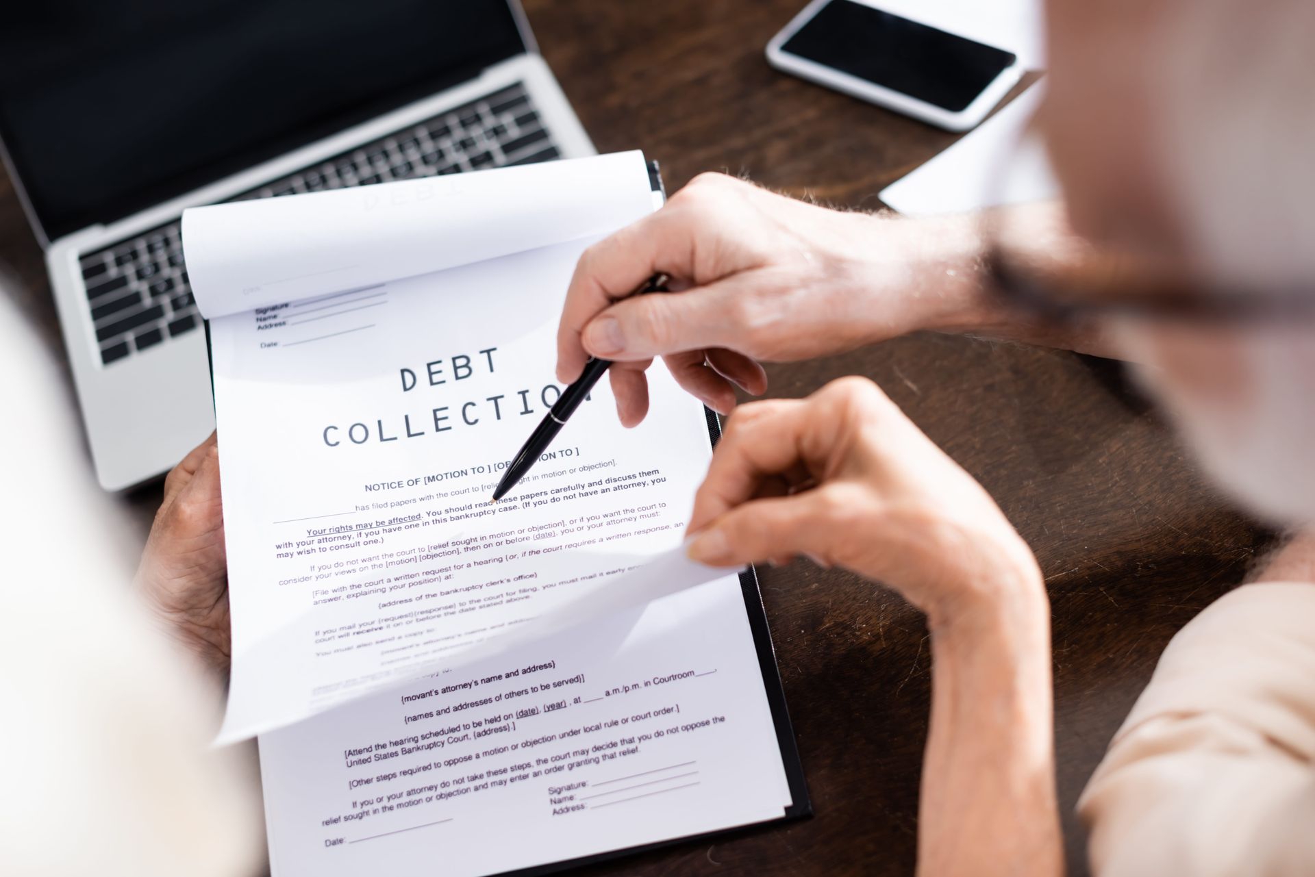 Hands reviewing a debt collection letter with a pen beside a laptop and phone on a desk