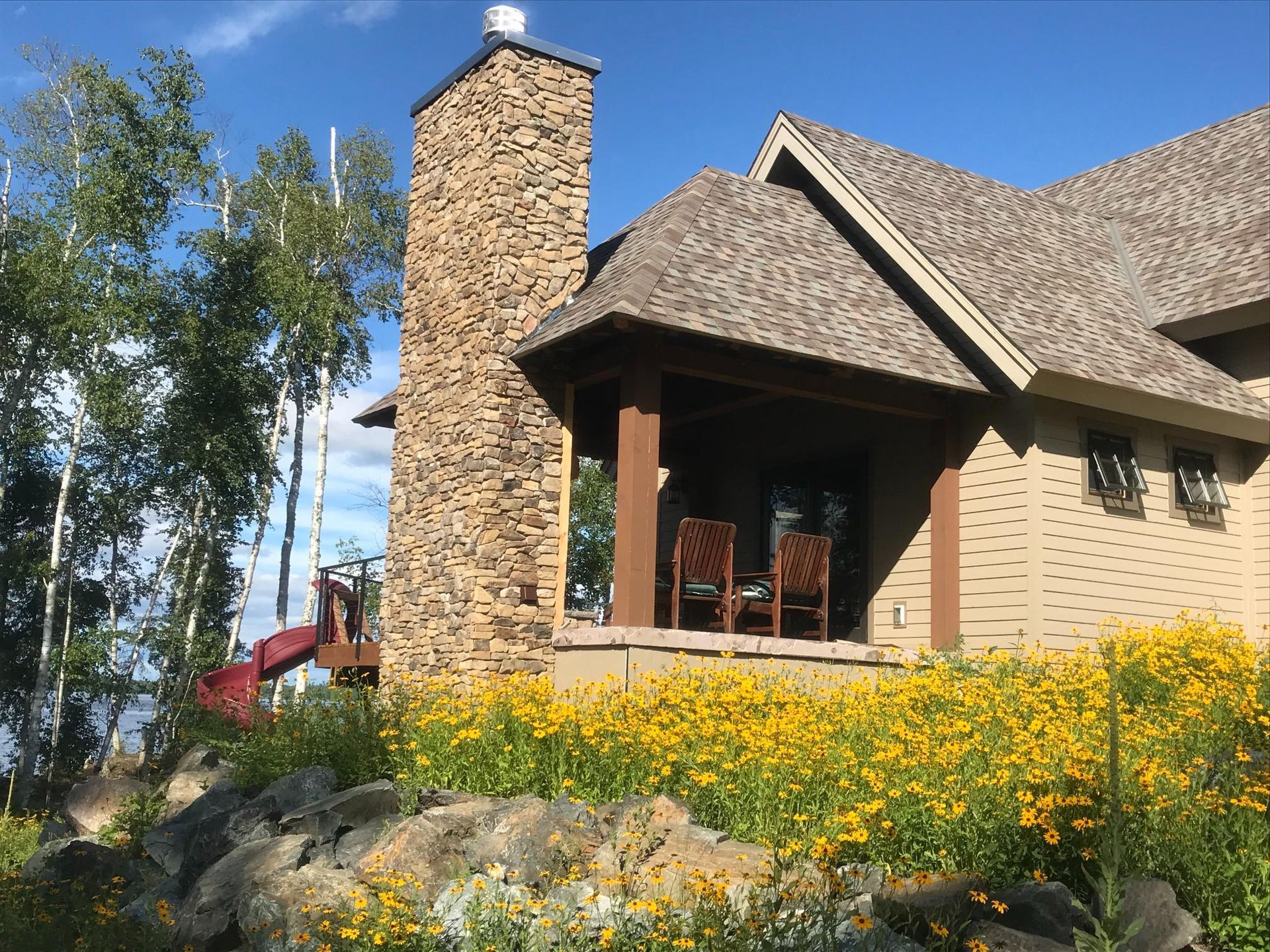 A house with a stone chimney is surrounded by yellow flowers and trees.