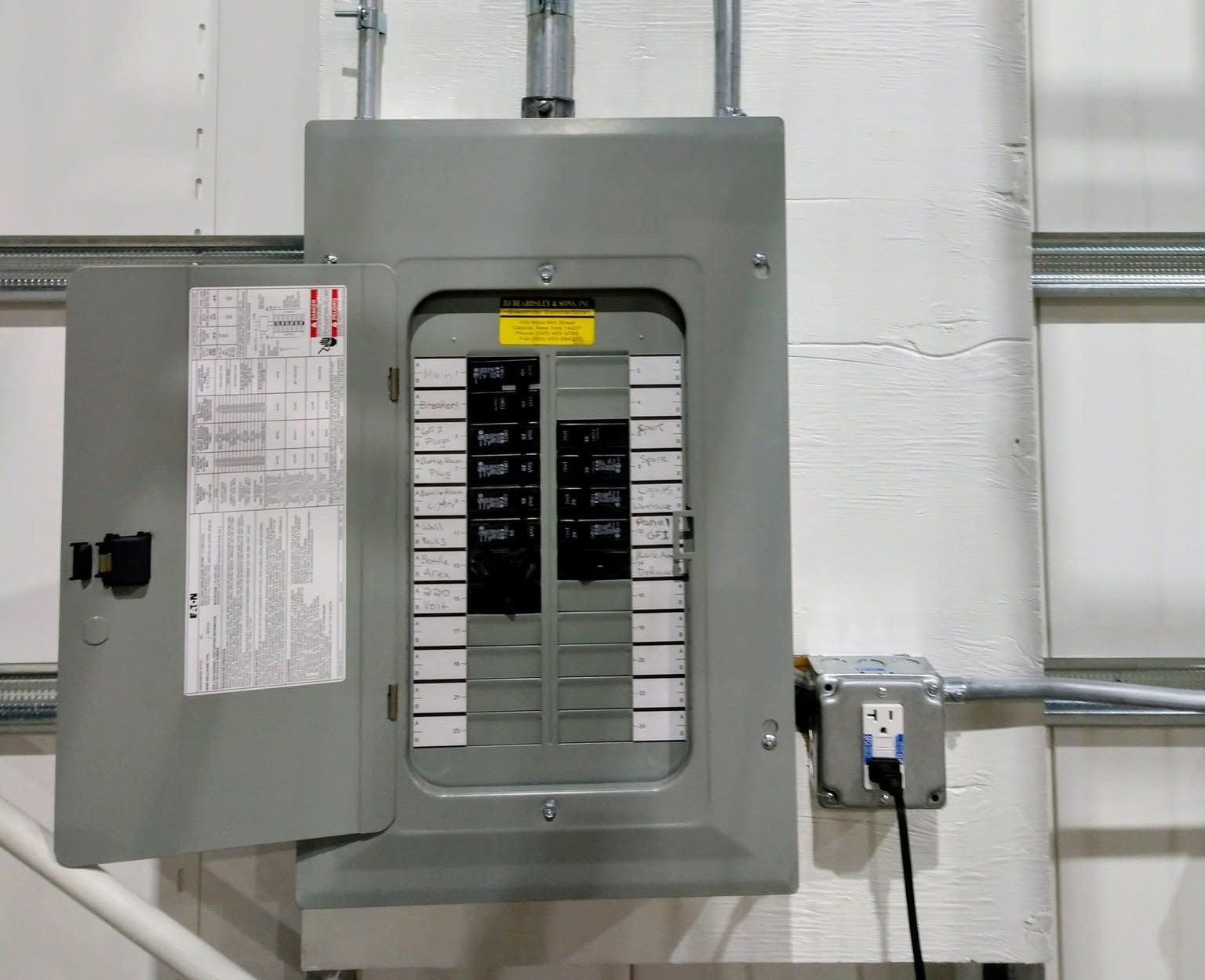 Open electrical breaker panel on a white wall with labeled switches and gray metal casing.