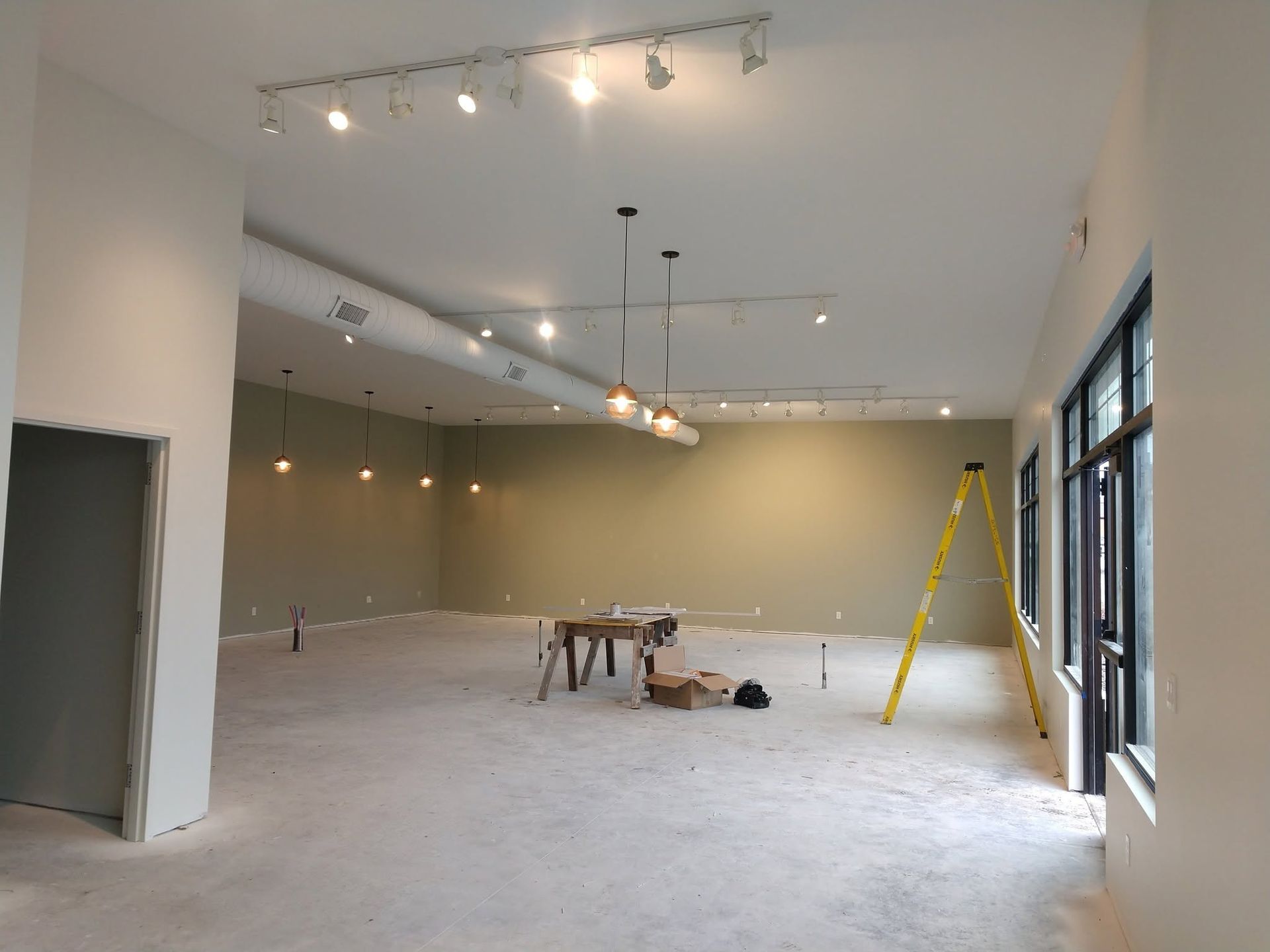 Empty commercial space with white walls, track lights, large windows, and a yellow ladder inside