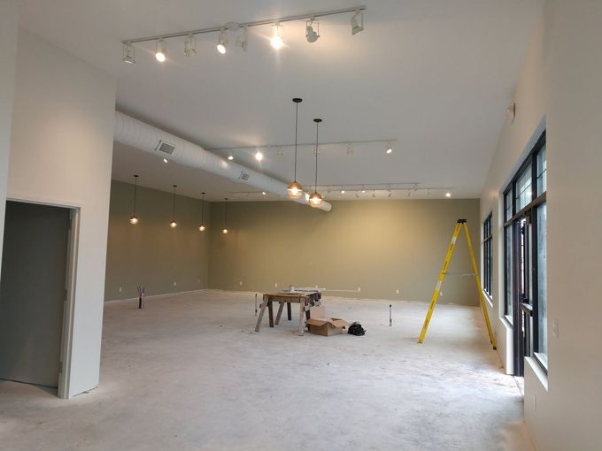 Empty room under construction with white walls, hanging lights, and a yellow ladder near large windows.