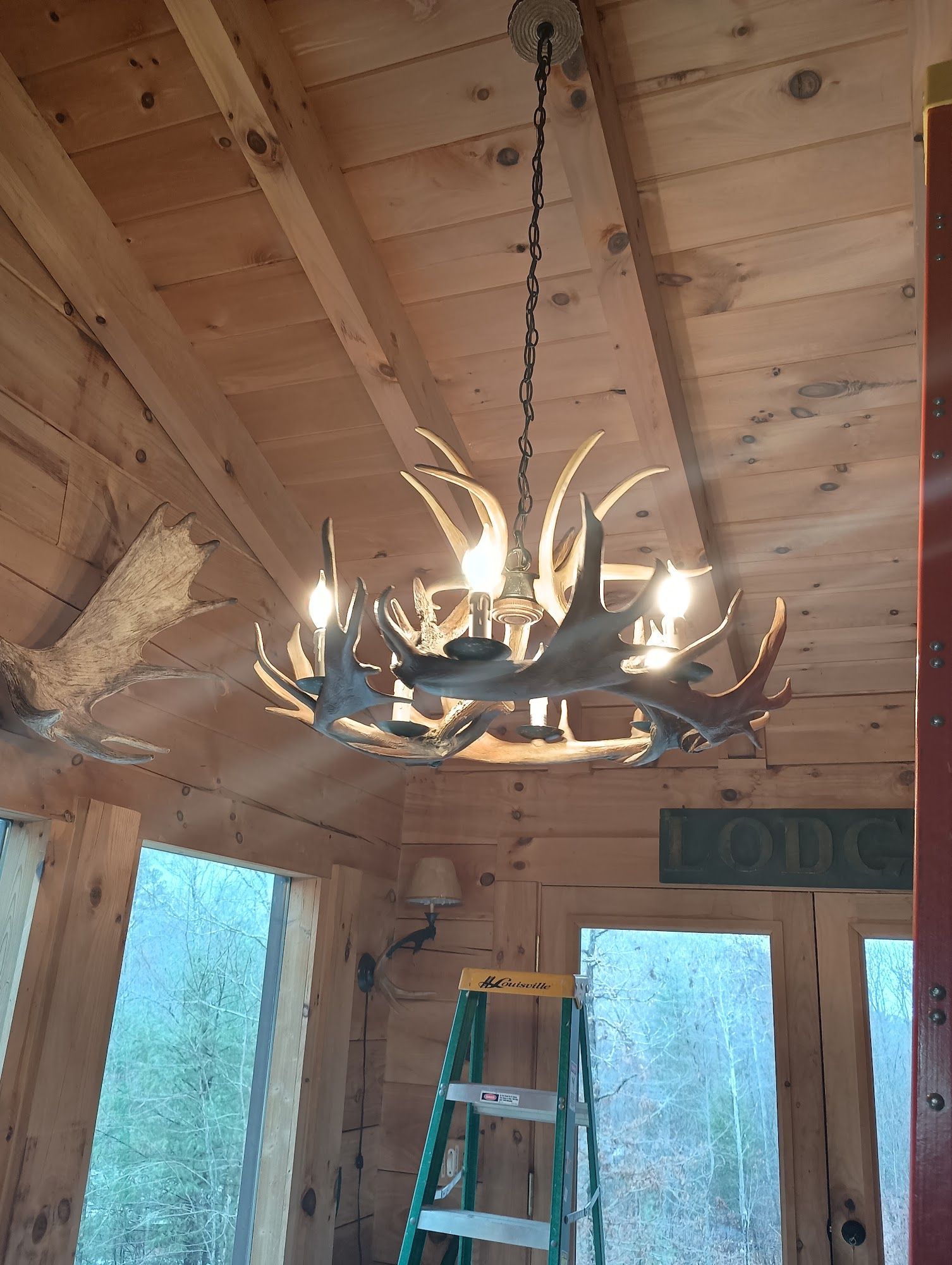 Rustic cabin interior with antler chandelier, ladder, wood-paneled ceiling, and windows.