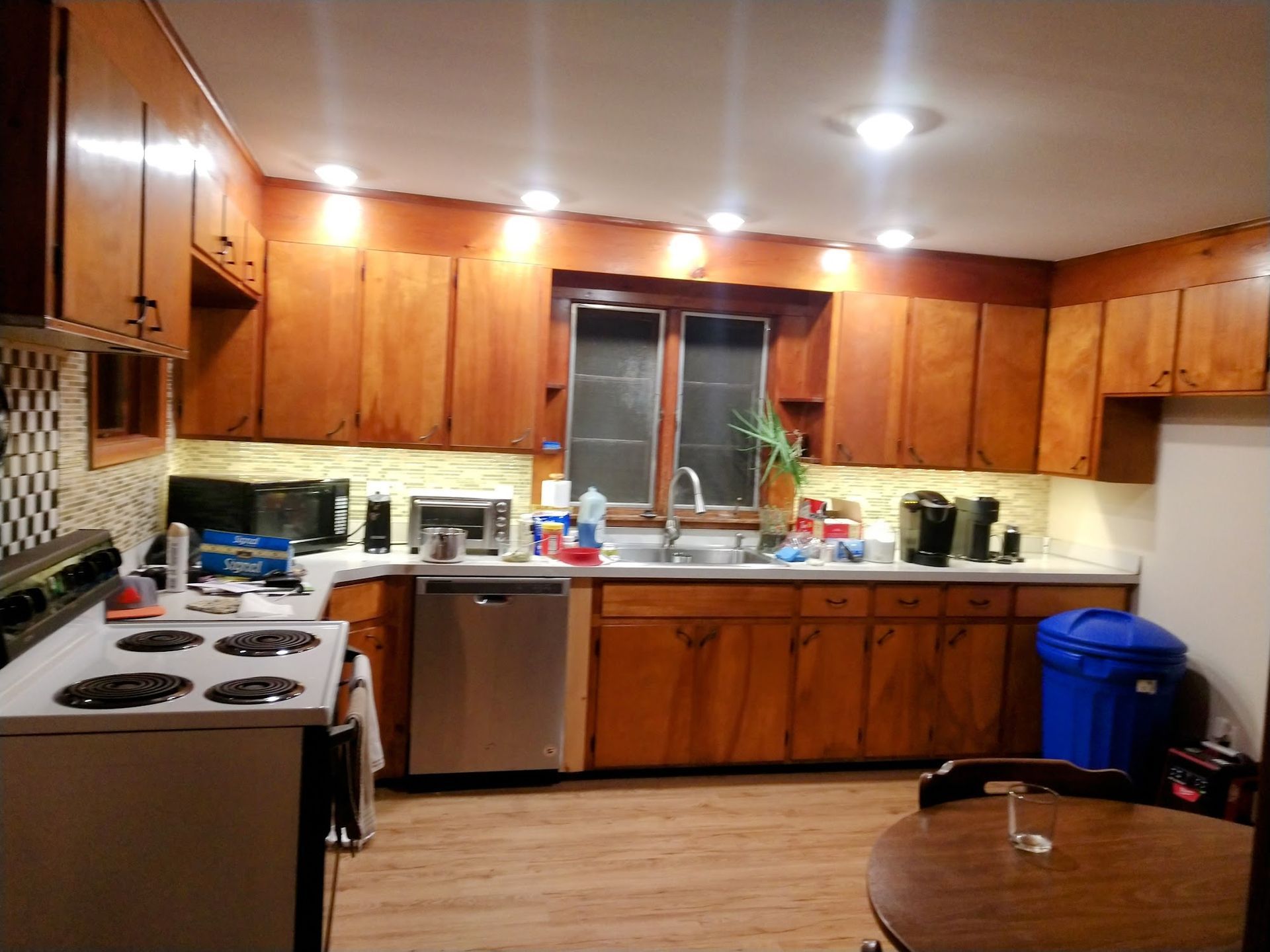 Warmly lit kitchen with wooden cabinets, white stove, sink by window, and blue recycling bin