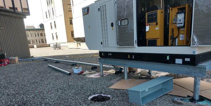 Outdoor industrial generator on gravel, with access panels open and cables on the ground.
