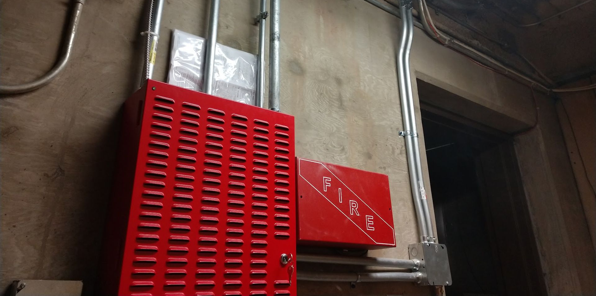 Red metal utility box and vented panel mounted on a wall beside silver pipes and a dark doorway.