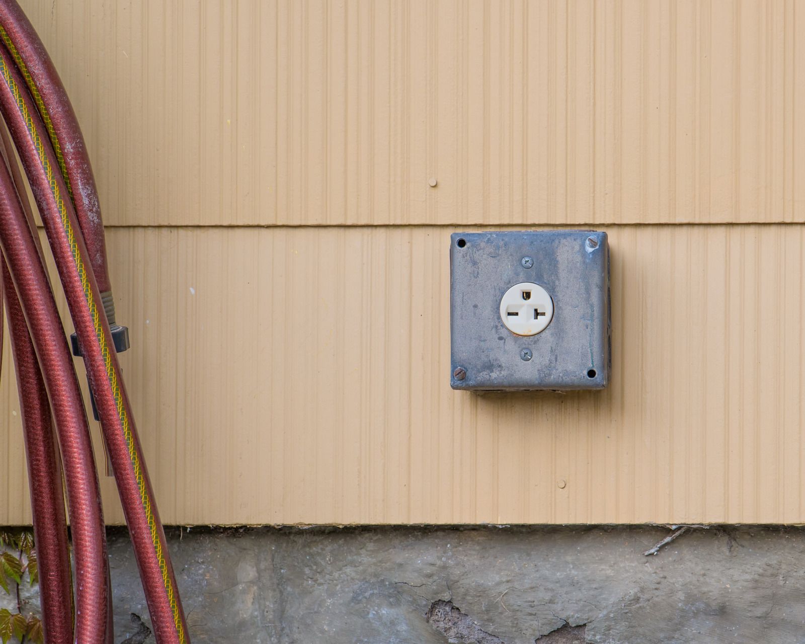 Exterior wall outlet in a metal box on beige siding, with coiled hoses along the left side.