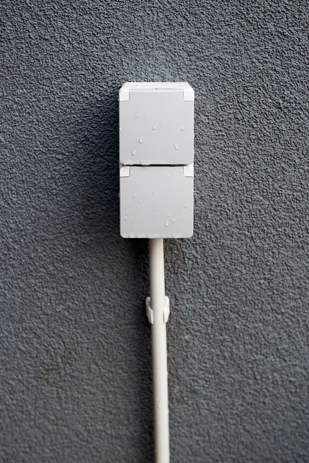 White electrical box on a gray textured wall with a conduit running downward
