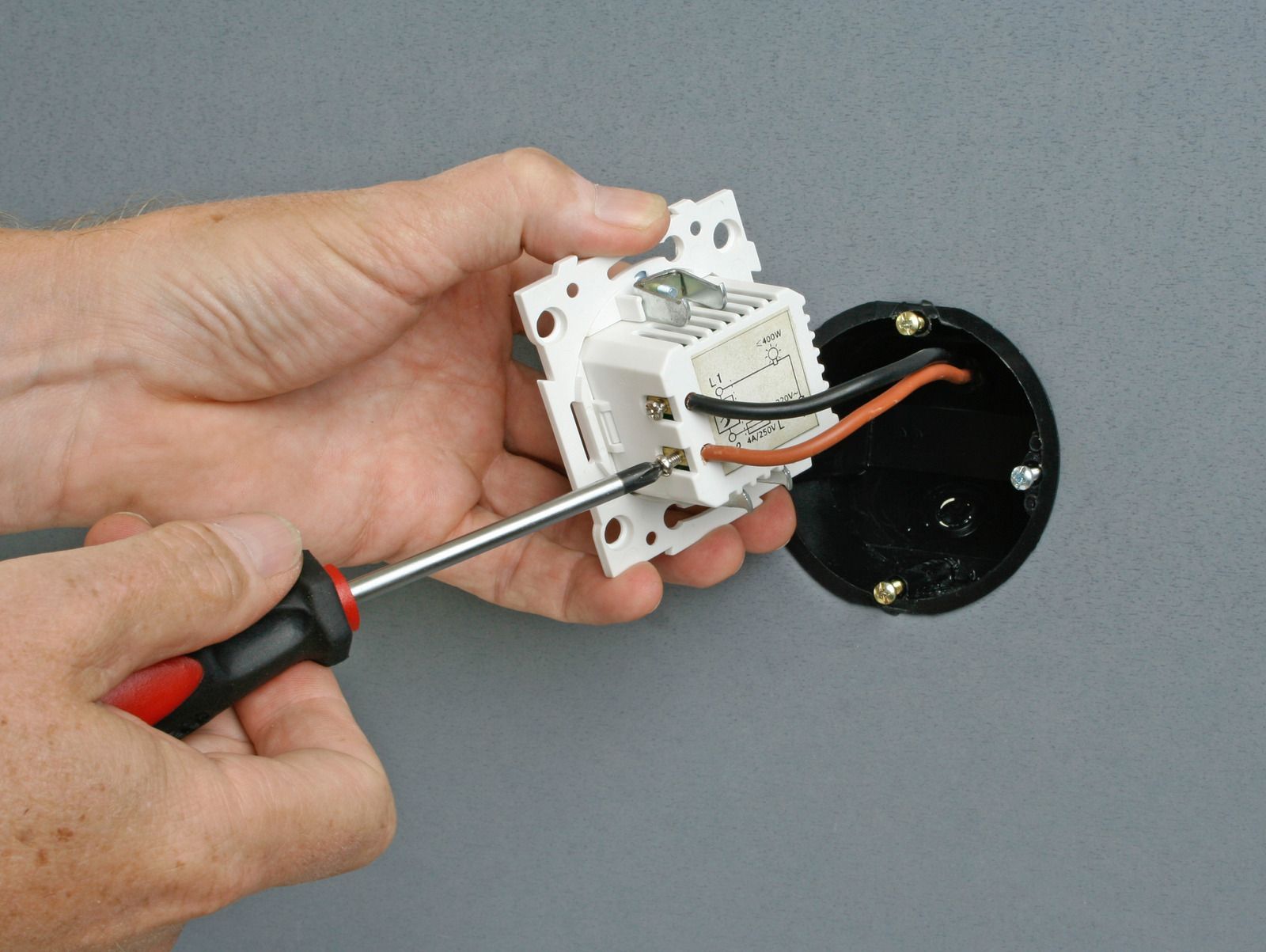 Hand using screwdriver to install a white electrical outlet on a gray wall, with black cable attached