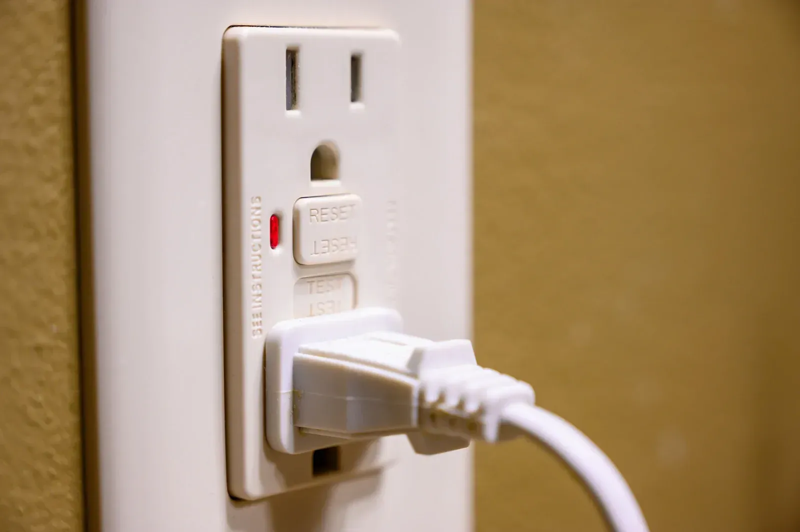 White wall outlet with a plugged-in power cord on a beige wall, showing a reset/test button.