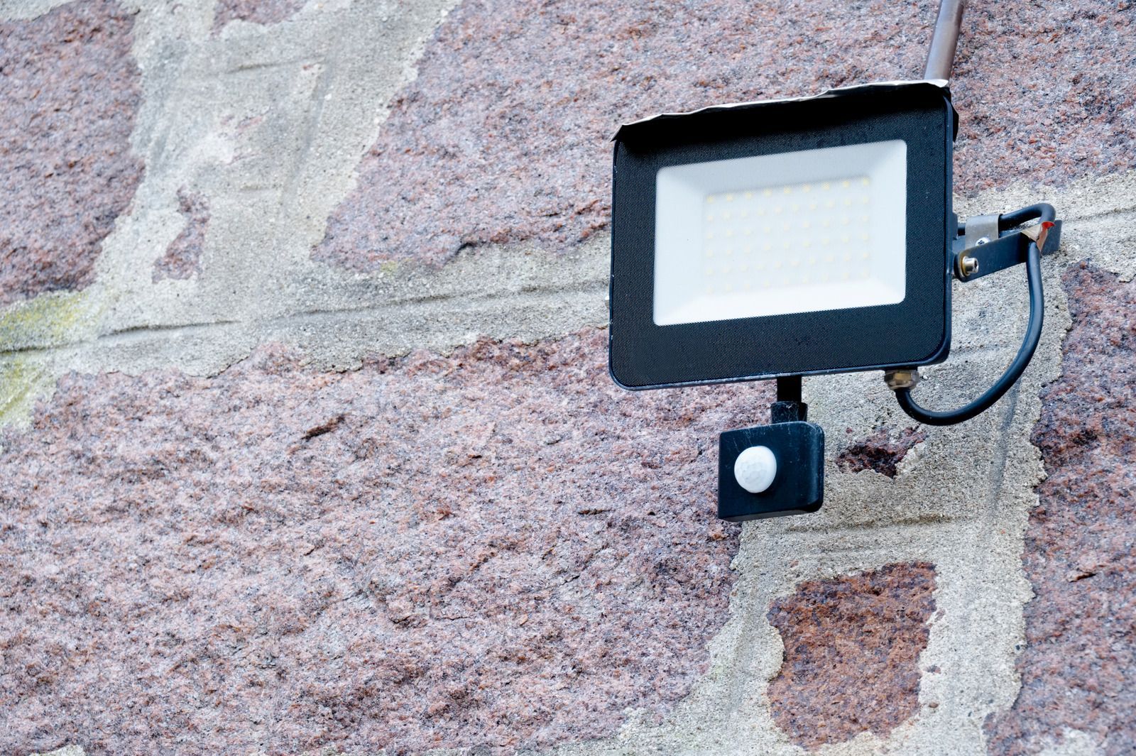 Black security floodlight mounted on a stone wall with a small motion sensor below it