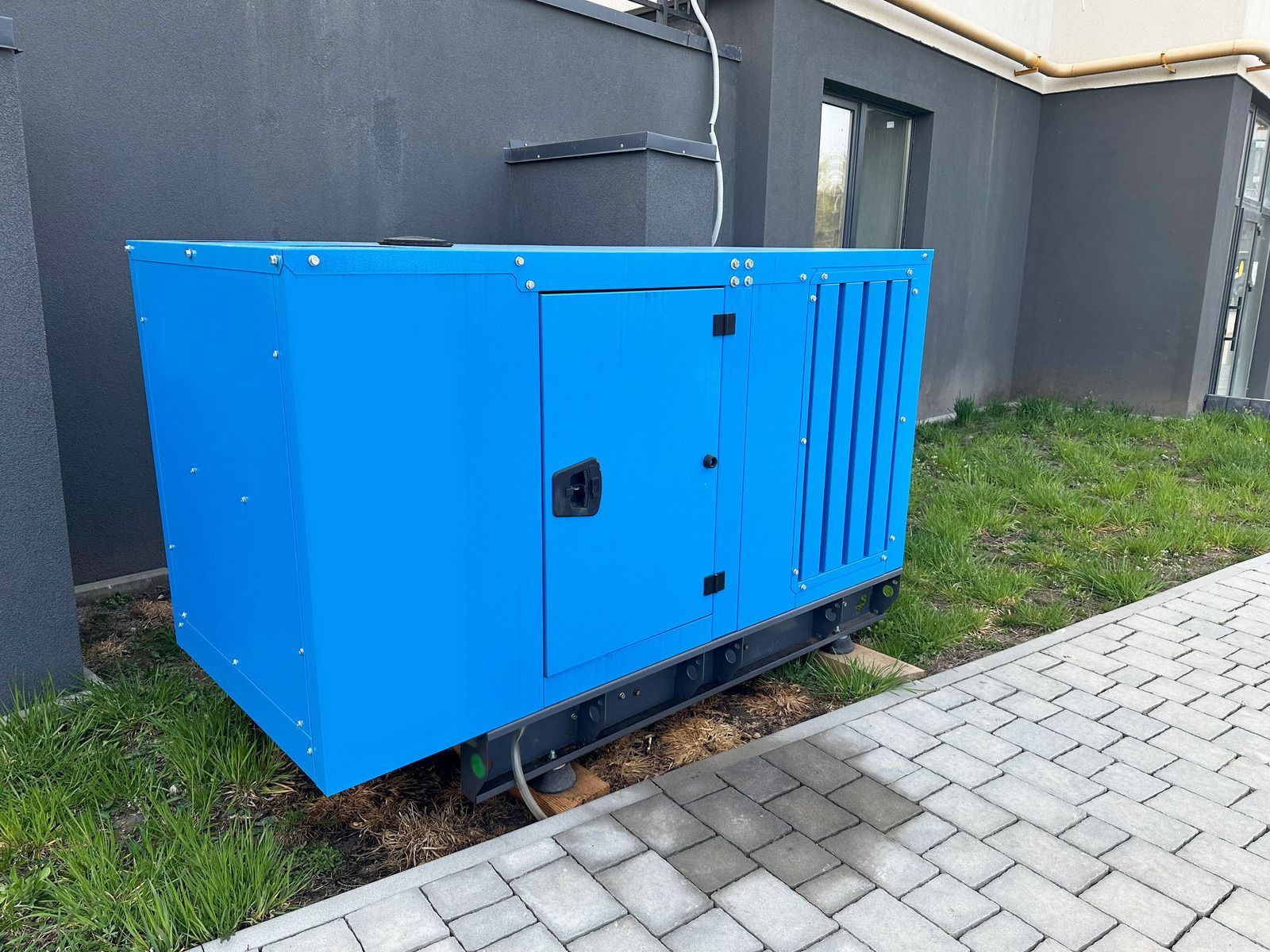 Blue industrial generator cabinet beside a building, next to grass and a paved walkway.