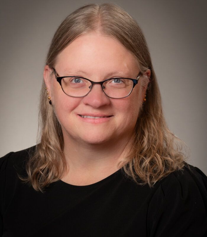 Woman with fair skin and glasses, smiling at the camera. She has blonde hair and wears a black shirt.
