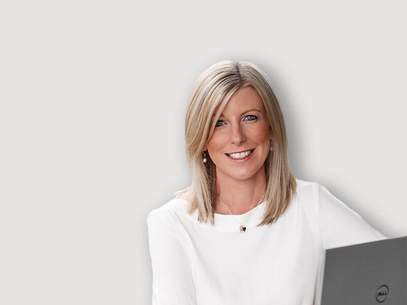 Woman smiling, in white top, holding laptop, against a light gray background.