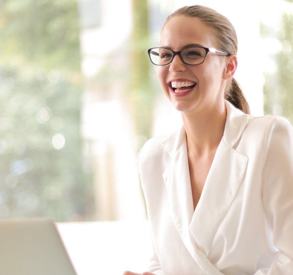 Woman in white blazer laughs, wearing glasses, near a laptop, with a bright window behind.