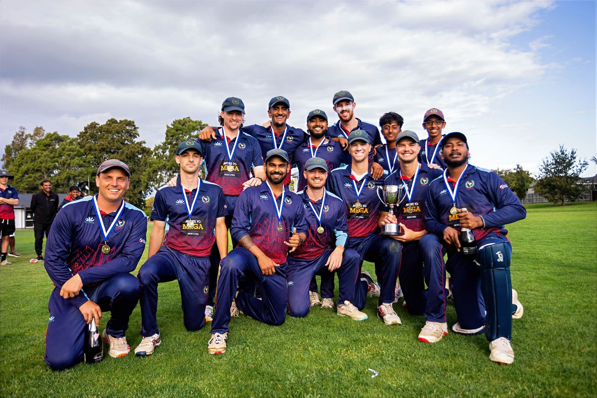Cricket team posing on a grassy field, wearing navy uniforms and medals, with trophies in front.