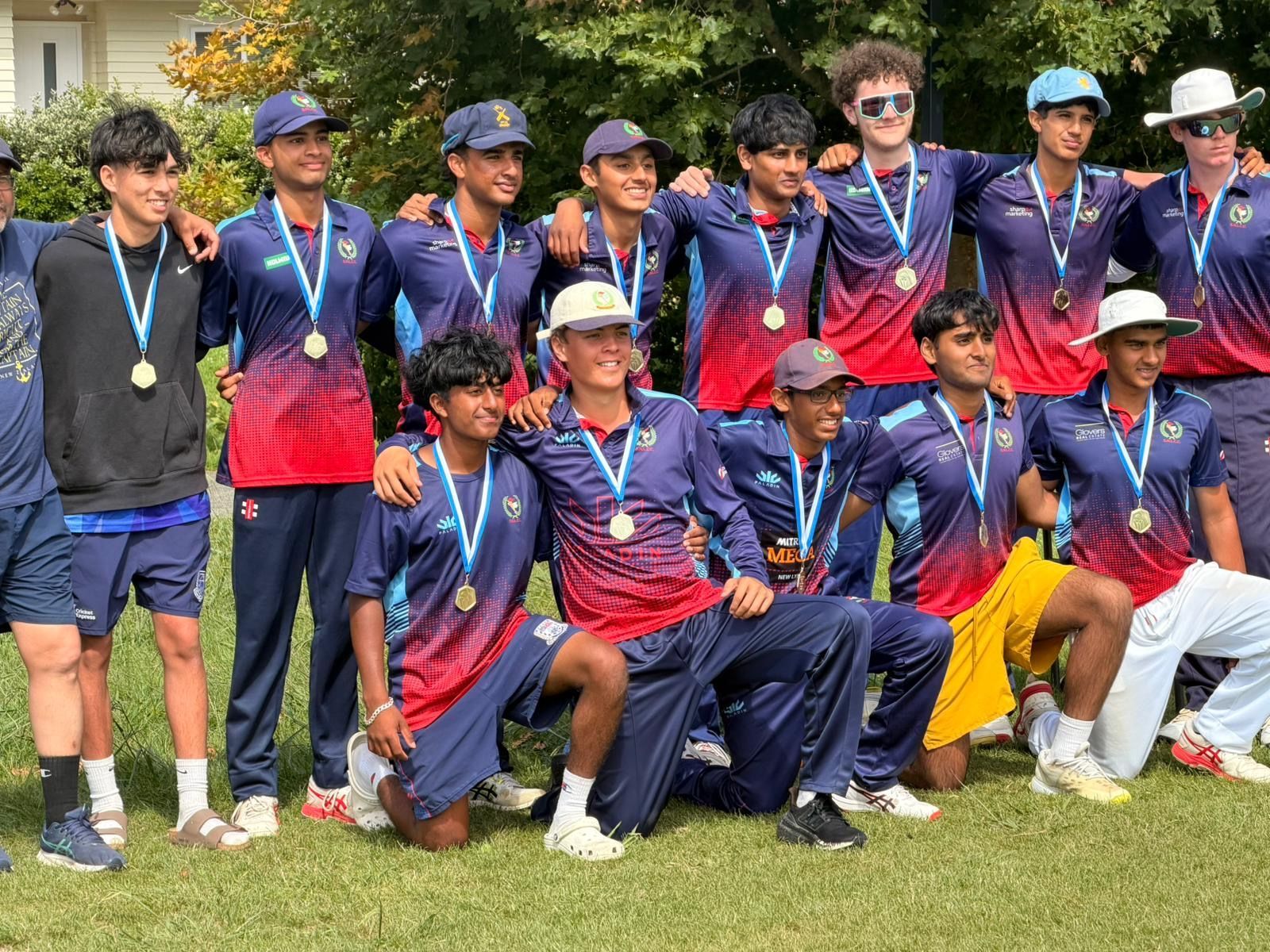 Group of cricket players in blue and purple uniforms posing on a grassy field with medals and trophies