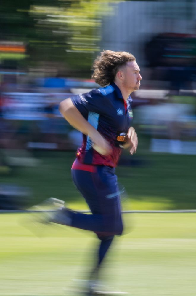 cricket bowler in navy and red sprinting outdoors on a blurred grassy course