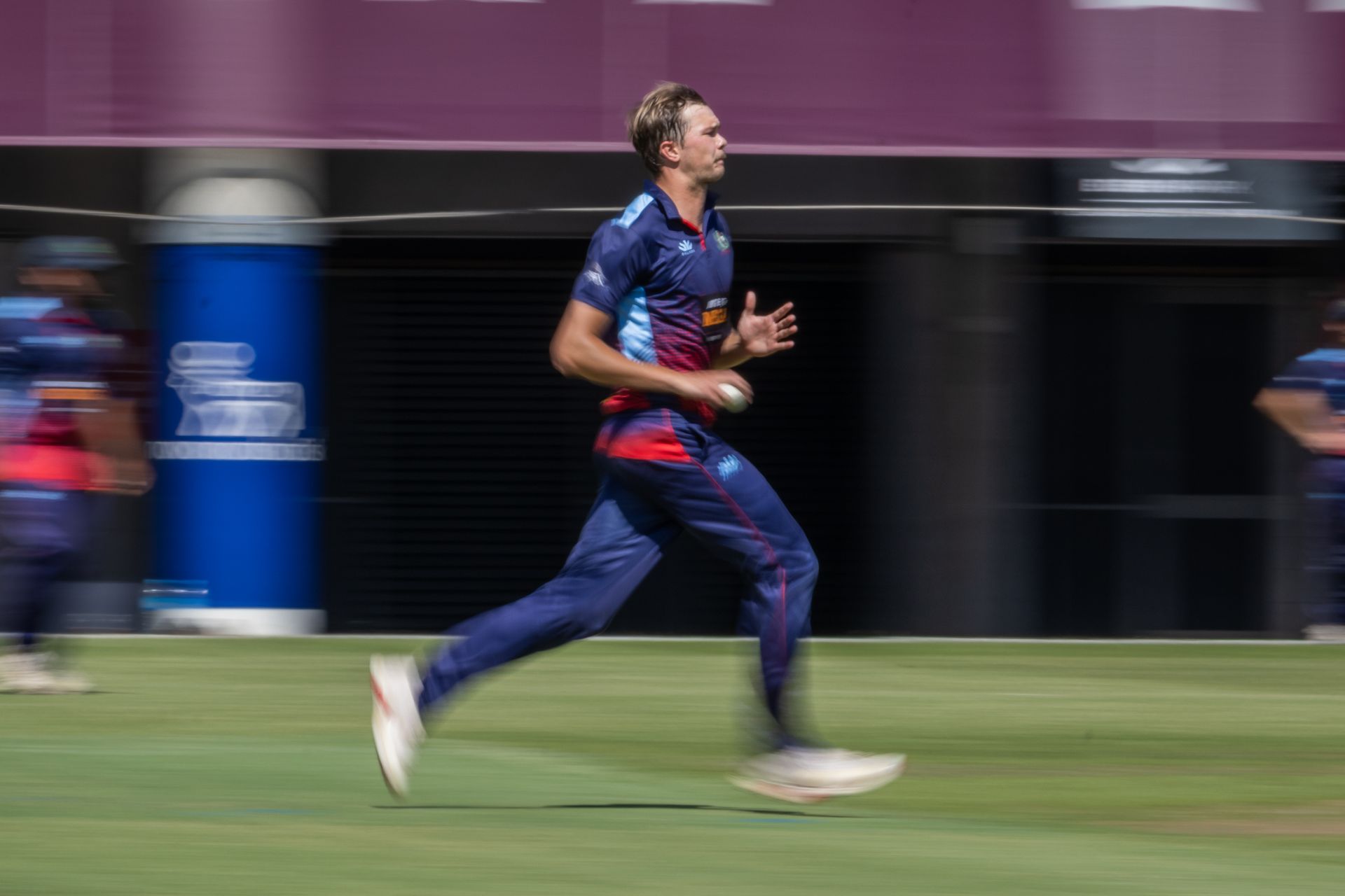 Cricketer in blue sprinting across the field during a match with a blurred stadium background