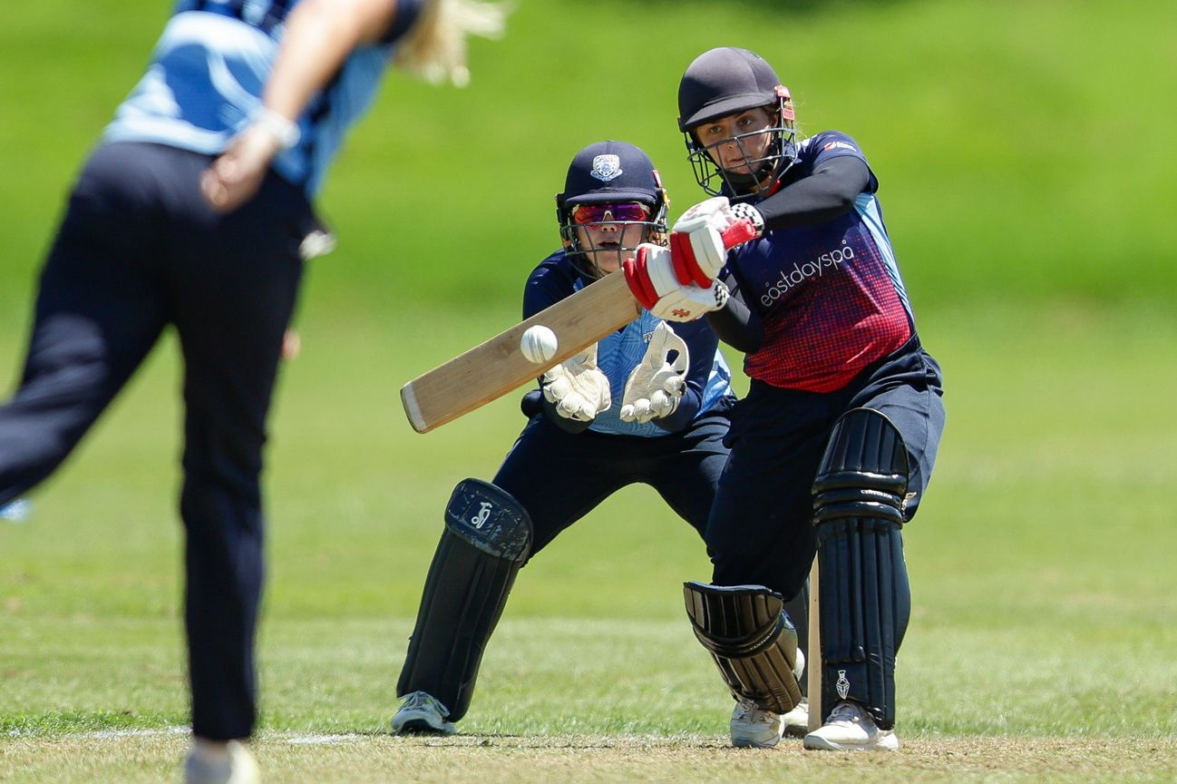 Cricket batsman in red and black swings at a ball while a wicketkeeper crouches behind him on a grassy field