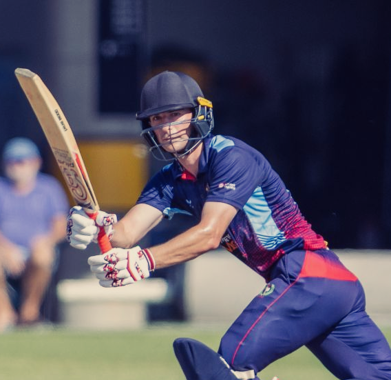 Cricket batter in navy and red uniform swings at the ball on a field