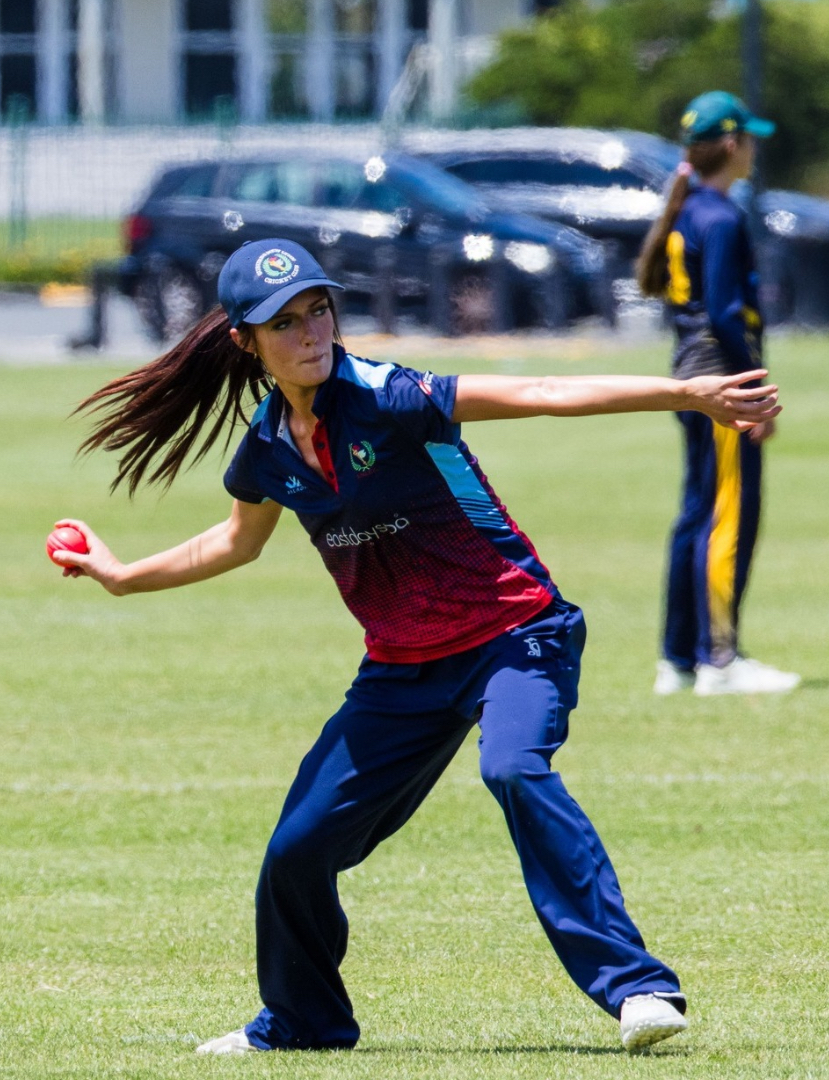 Cricketer in navy and maroon uniform bowling on a grassy field, with a teammate in the background.
