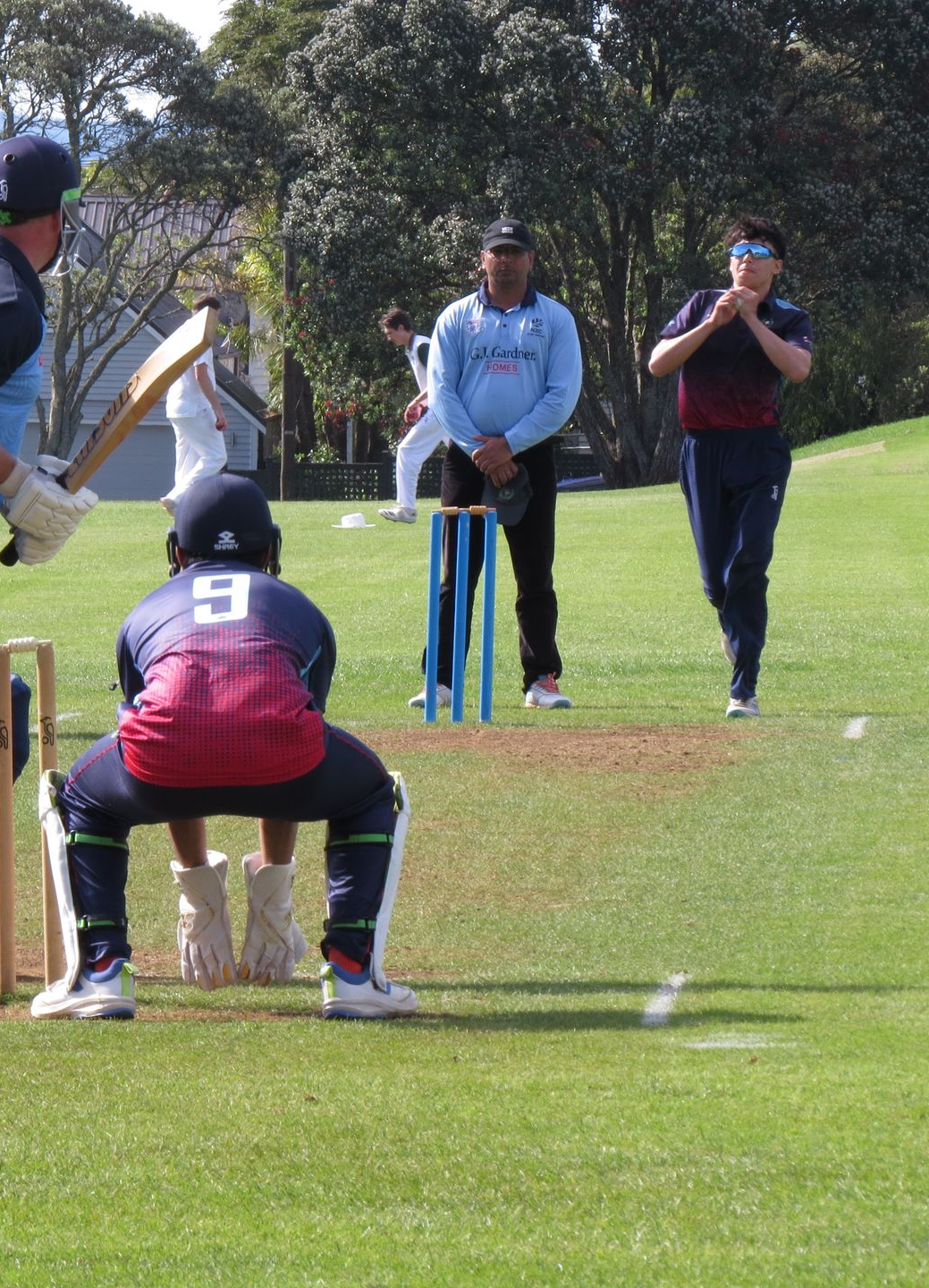 Cricket batsman crouches near wicket as bowler and fielders stand on a grassy pitch.