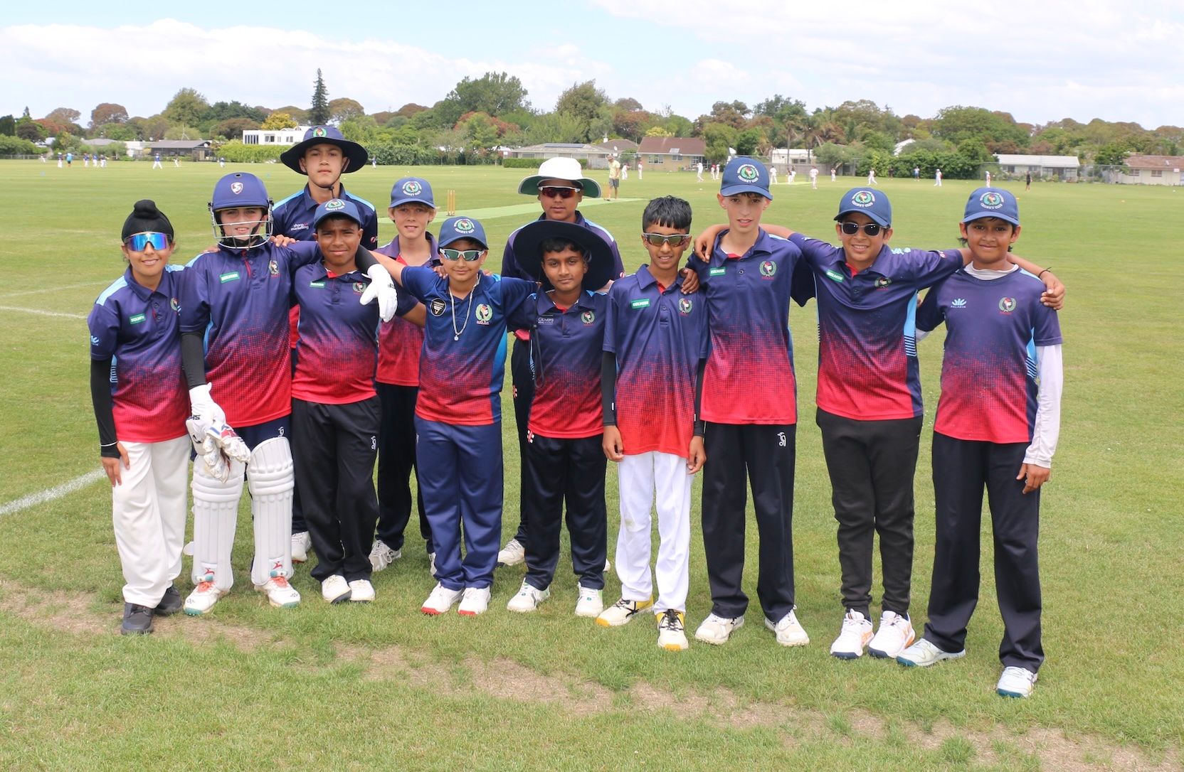Youth cricket team posing on a grassy field in blue, red, and navy uniforms with bats and helmets