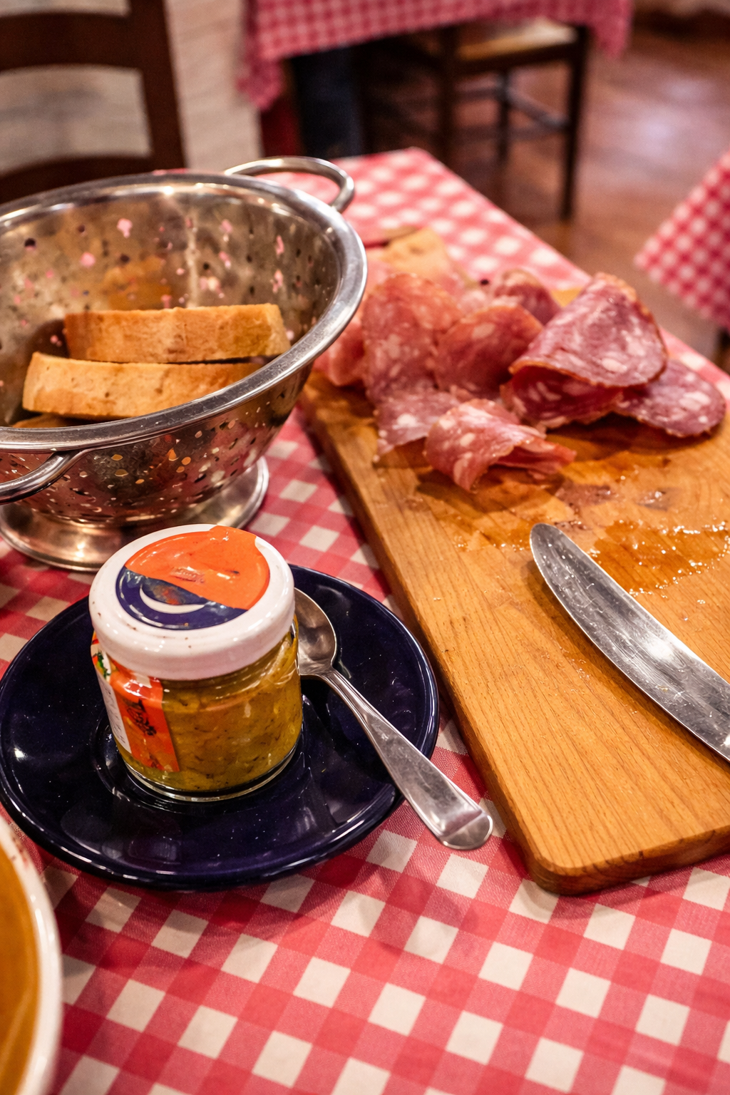 Tavola apparecchiata con pane, salame su un tagliere, barattolo di senape e un cucchiaio. Tovaglia a quadri rossi.