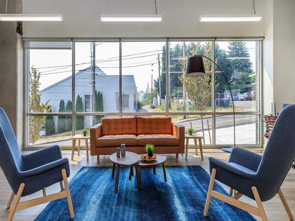 Lounge area with orange sofa, two blue chairs, and a blue rug in front of large windows.
