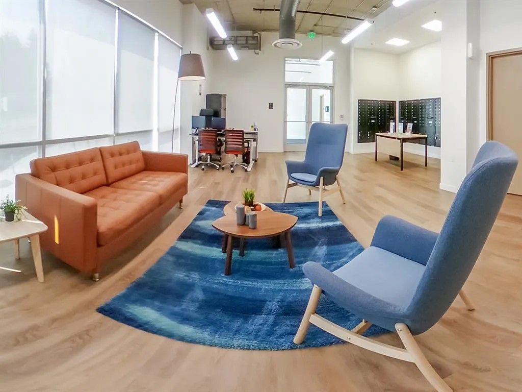 Lounge area with orange sofa, blue chairs, rug, round table, and a desk in the background.