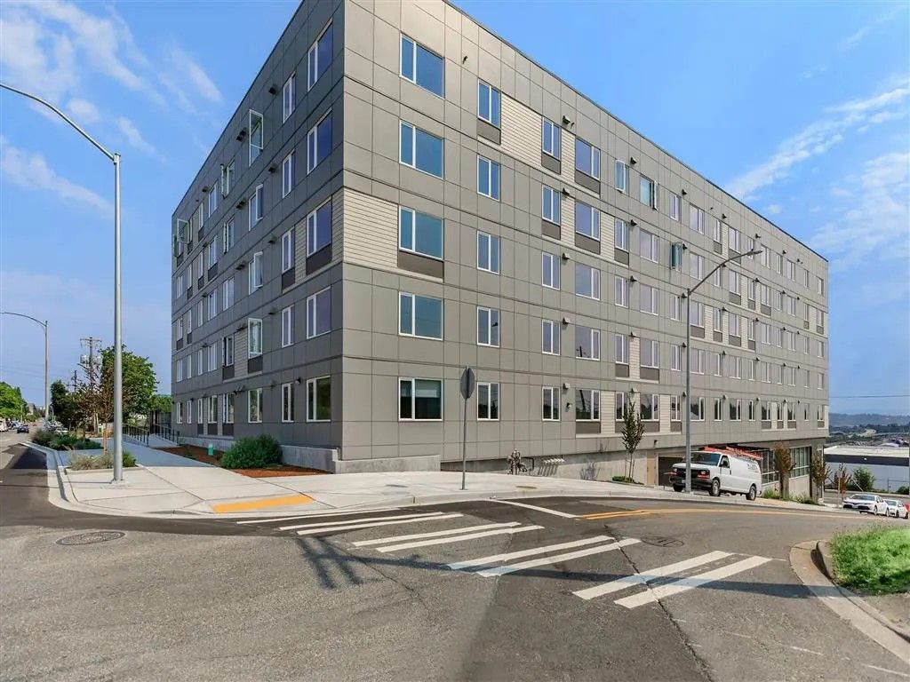 Gray multi-story building on a city street, crosswalk in foreground, blue sky.