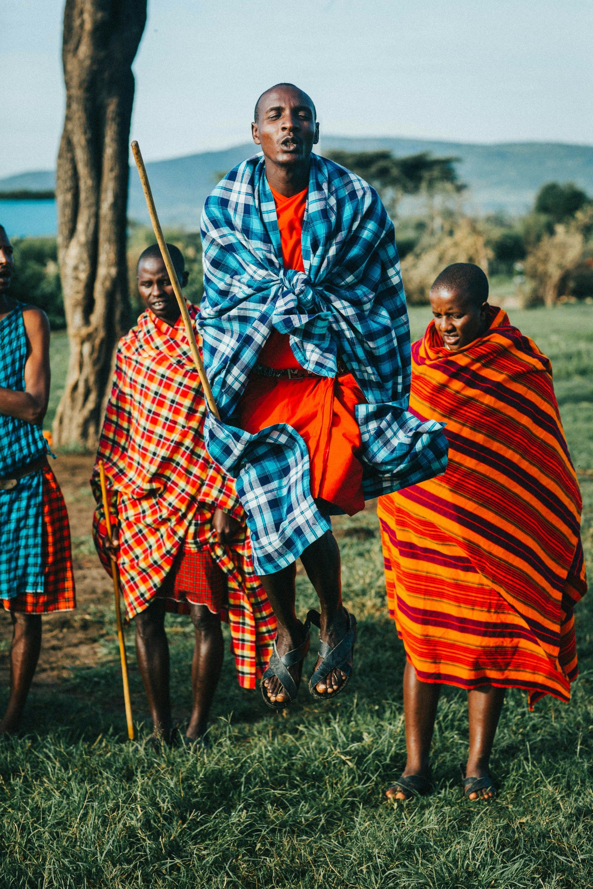 People in traditional colorful shuka wraps dancing in a grassy field, with one person leaping mid-air.