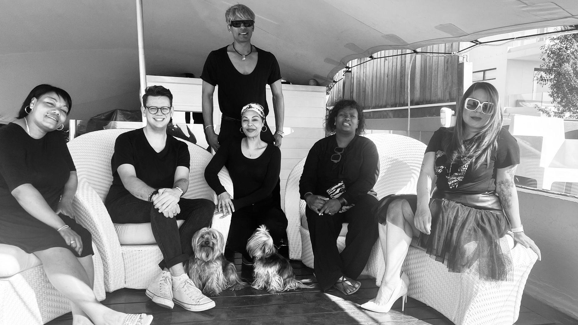 A group of six people posing together on light-colored outdoor lounge furniture, accompanied by two small dogs.