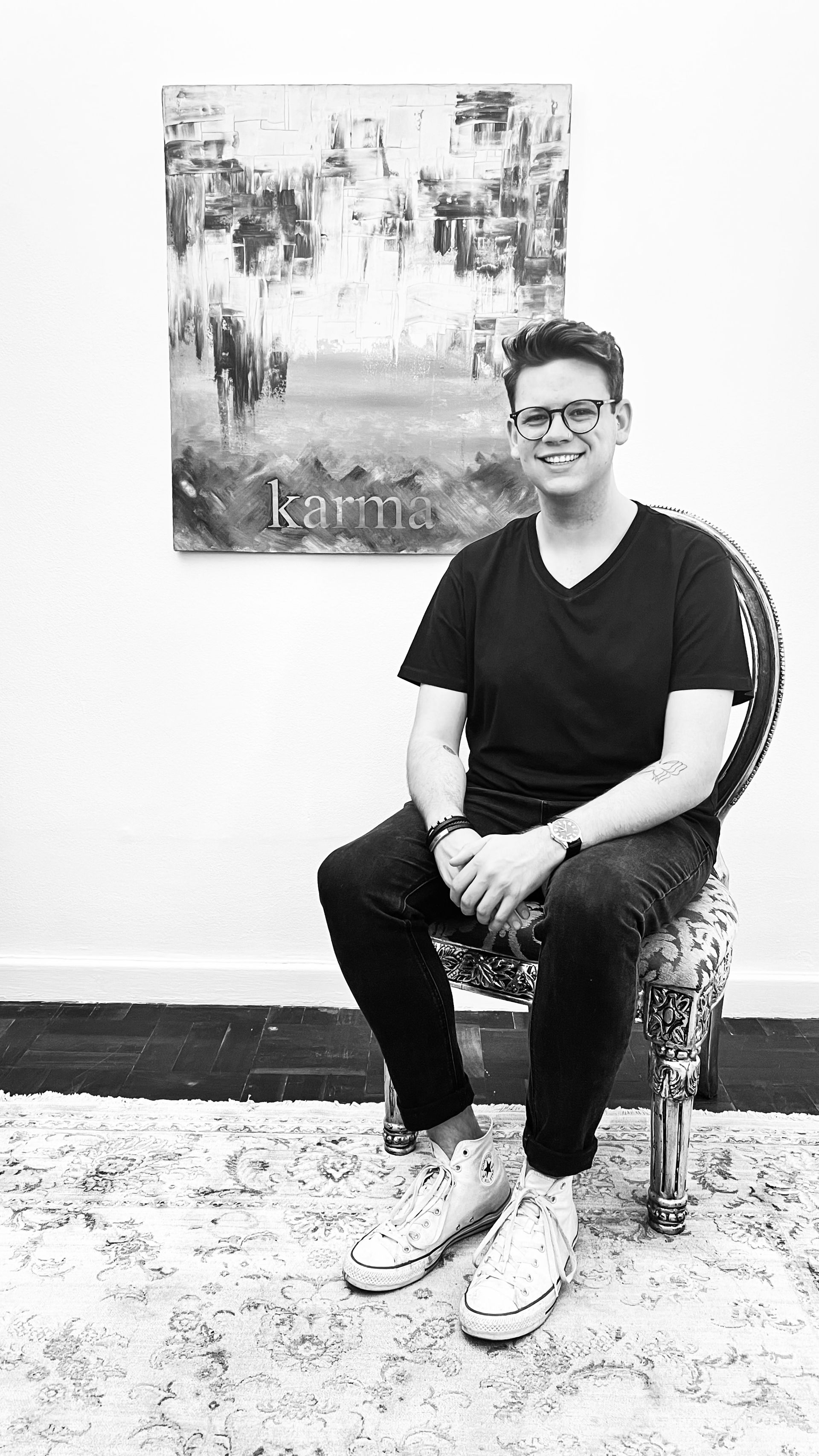 A person wearing glasses and a black shirt sits on an ornate chair in front of a framed painting against a white wall.