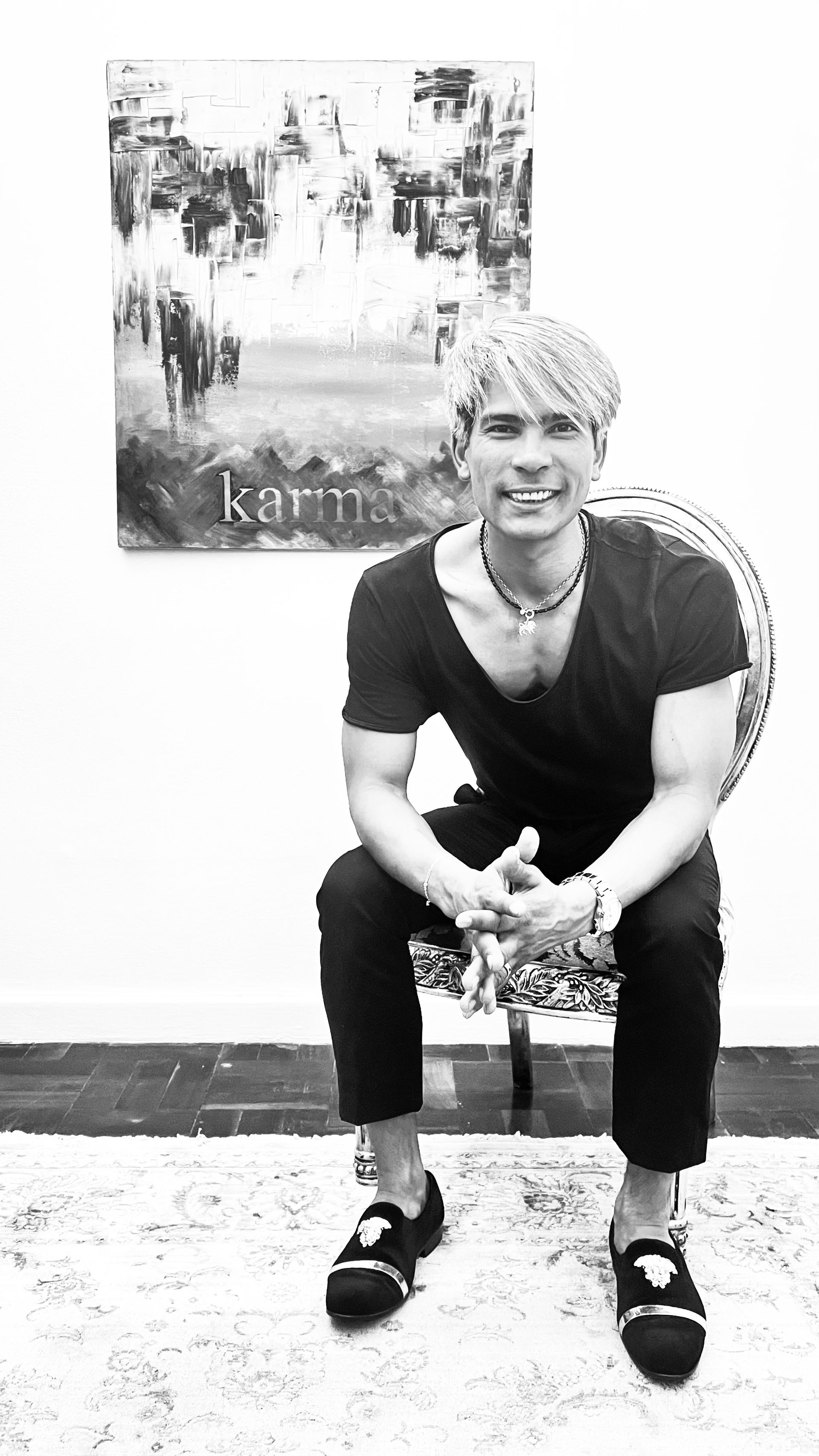 A person with light-colored hair smiles while sitting in a patterned chair before a wall art piece with the word 
