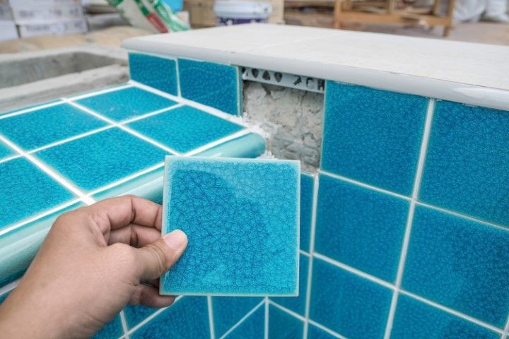 Hand holding a blue tile near a partially tiled wall during tile installation