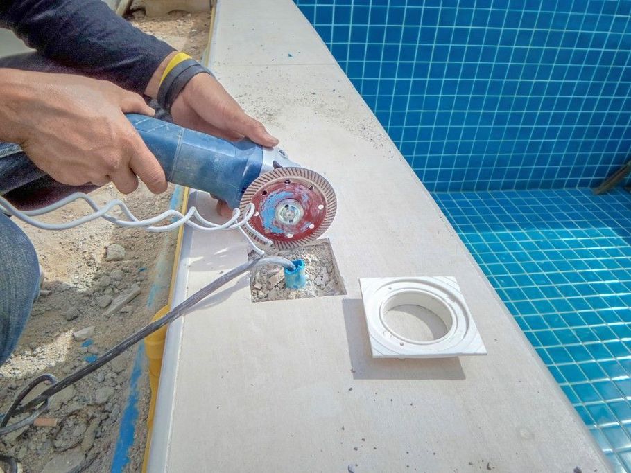 Worker using a grinder to cut tile beside a blue-tiled pool deck, with a drain opening nearby